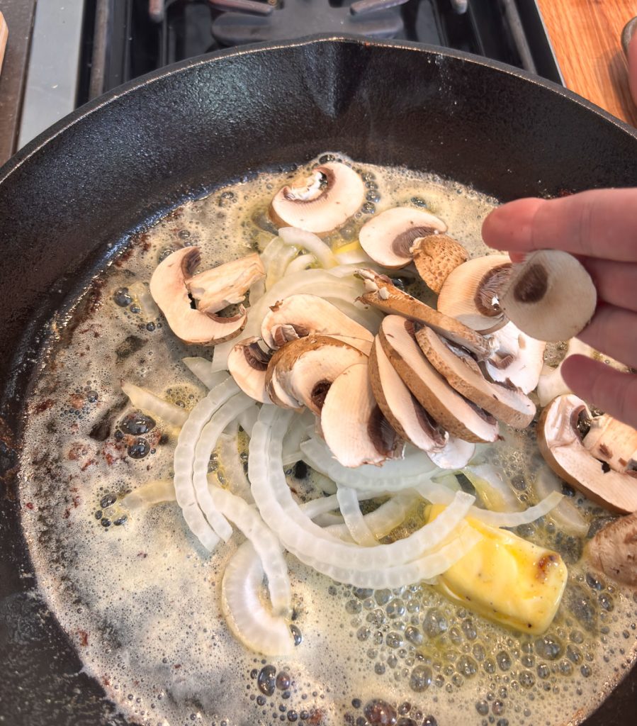 melted butter in a cast iron skillet with a hand coming in from the right adding sliced onions and white button mushrooms to the pan