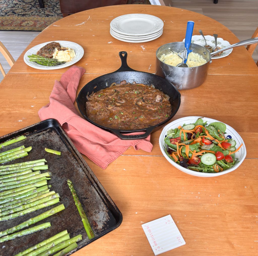 dinner table with skill of swiss steak, sheet pan of asparagus, a green salad, a pan of mashed potatoes, and table settings, nothing fancy