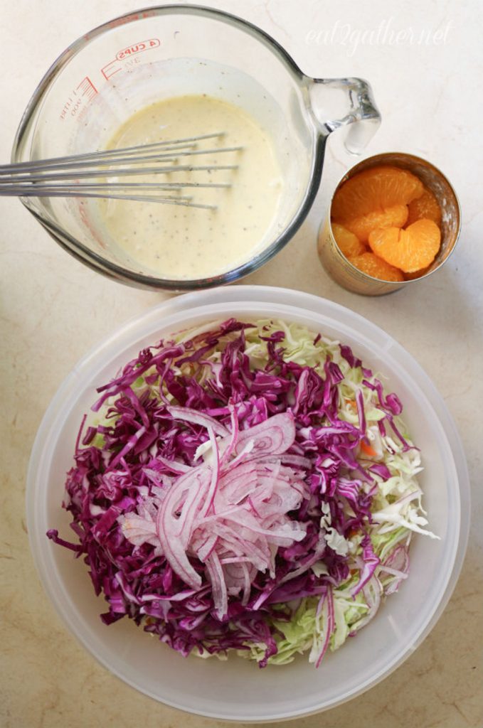 glass bowl of slaw dressing in upper left corner, open can of mandarin oranges in upper right, bowl of salad ingredients in bottom half of the photo all on a white background