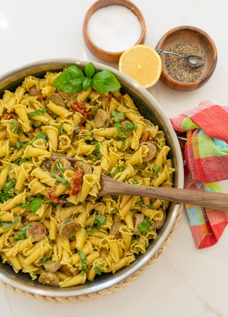 pan of sundried tomato and pesto pasta on a white countertop with a wooden spoon sitting in the pan. wood bowls of salt and pepper and a half lemon are next to the pan