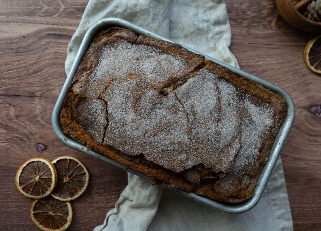oaf of pumpkin bread in bread pan on a wood counter top with a white and tan gingham tea towel and slices of dried orange