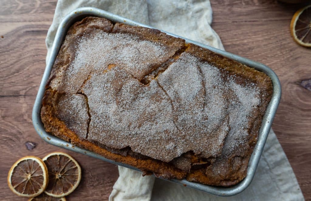 loaf of pumpkin bread in bread pan on a wood counter top with a white and tan gingham tea towel and slices of dried orange
