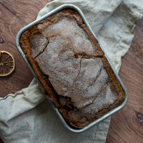 loaf of pumpkin bread in bread pan on a wood counter top with a white and tan gingham tea towel and slices of dried orange