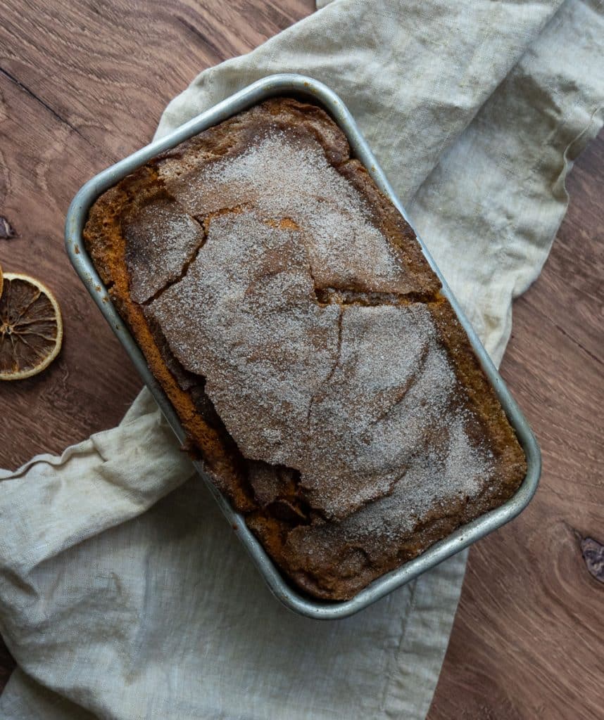 loaf of pumpkin bread in bread pan on a wood counter top with a white and tan gingham tea towel and slices of dried orange