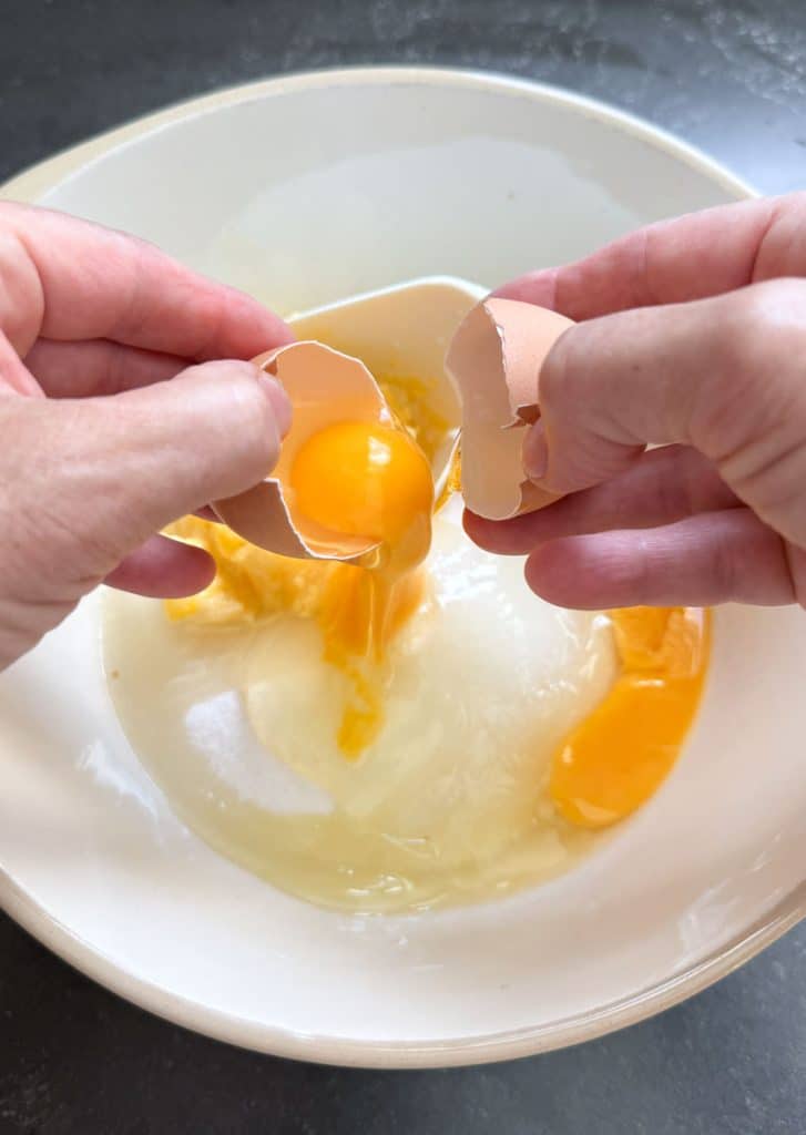 Cracking eggs into white mixing bowl with granulated sugar in it
