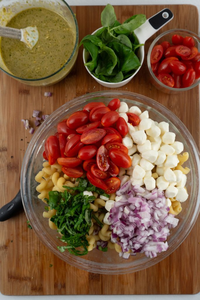 glass bowl full of noodles, tomatoes, basil, onion, mozzarella pearls, on top of wood cutting board with dressing, basil and tomatoes above the bowl 