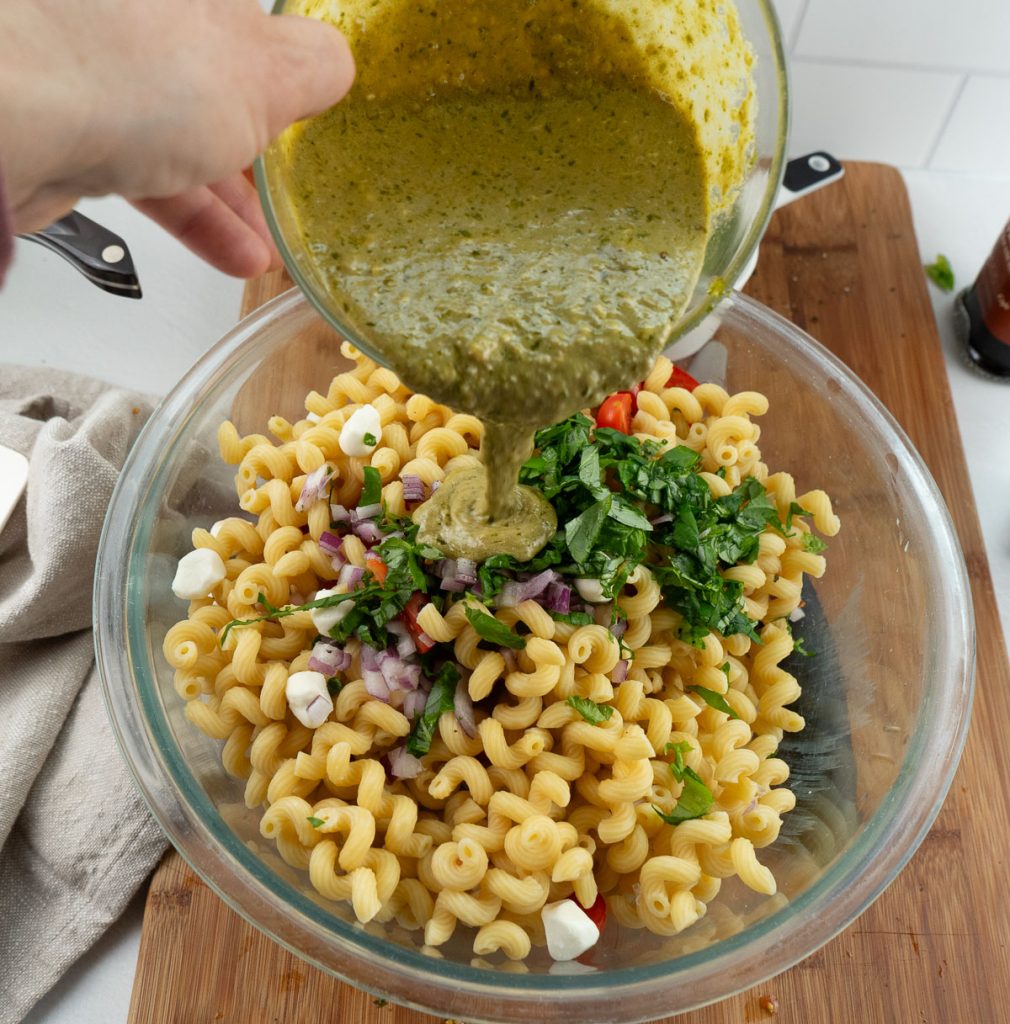 glass bowl full of pasta salad ingredients with dressing being poured over it