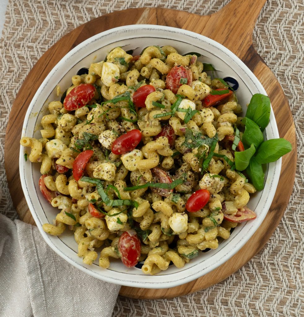 white bowl full of pesto caprese pasta salad on a round wood cutting board with tan background