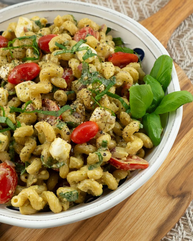 noodles, tomatoes, basil, mozzarella pasta salad in a white bowl on a wood cutting board 