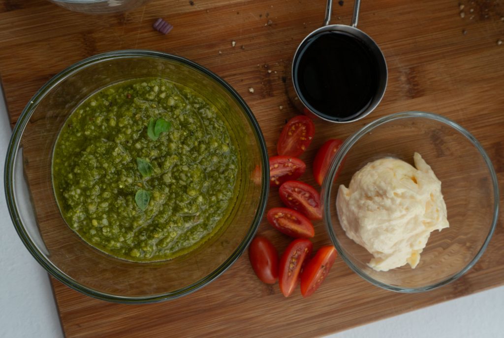 pesto, balsamic vinegar, mayonnaise in glass bowls on a wood cutting board with some cherry tomatoes scattered on the board