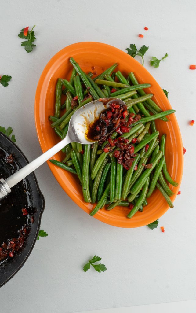 Green beans, on a orange platter with sweet and sour bacon sauce being spooned over green beans