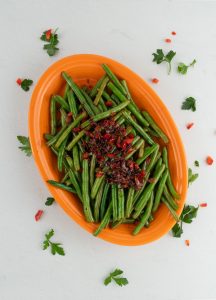 Green beans on a orange platter with bacon and brown sugar glaze on top with sprinkles of red pepper and parsley.