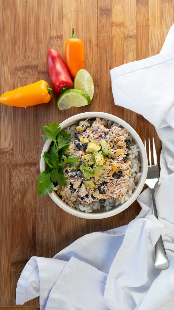 white bowl with rice and creamy black bean andc chicken with green garnish sitting on a wood cutting board with peppers and limes. 