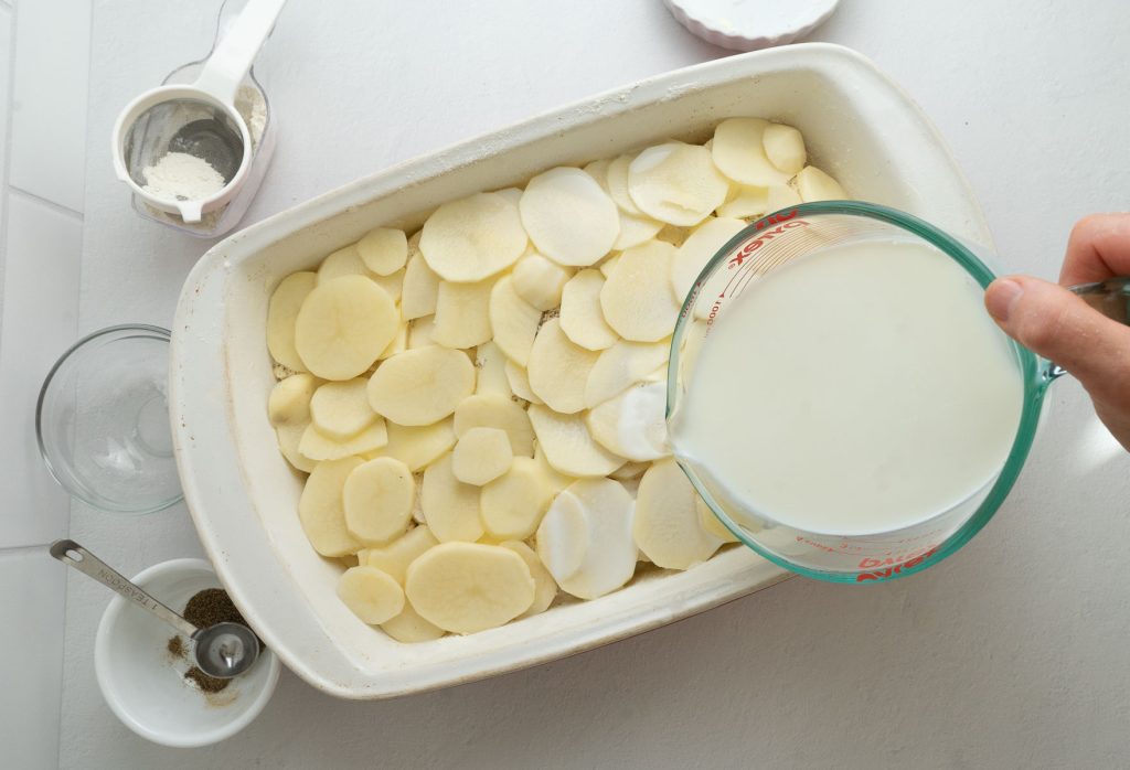 potatoes layered in a casserole dish with warm milk being poured over them from a measuring cup
