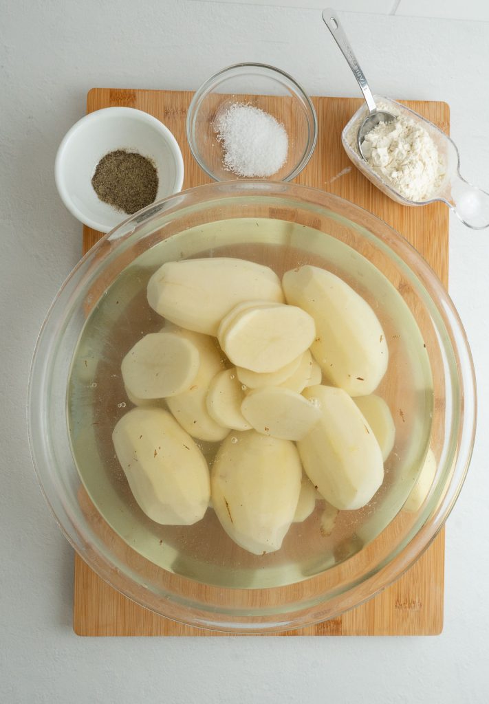 bowl of water with peeled potatoes in it sitting on a wood cutting board. salt pepper and flour are in small bowls at top