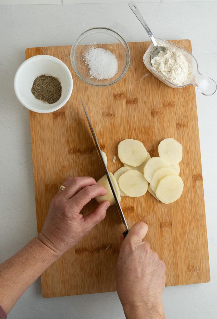 slicing peeled potatoes with a knife on a wood cutting board dishes of salt pepper and flour to the side