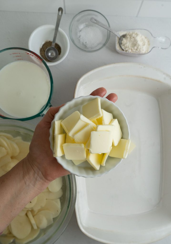 scallop potato ingredients in background with a hand holding a dish of butter slices in fore front 