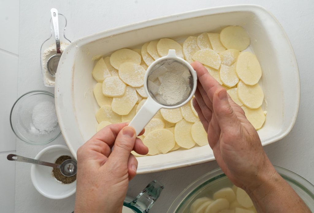 process photo for scallop potato recipe. layer of potatoes in dish sprinkling flour over top 