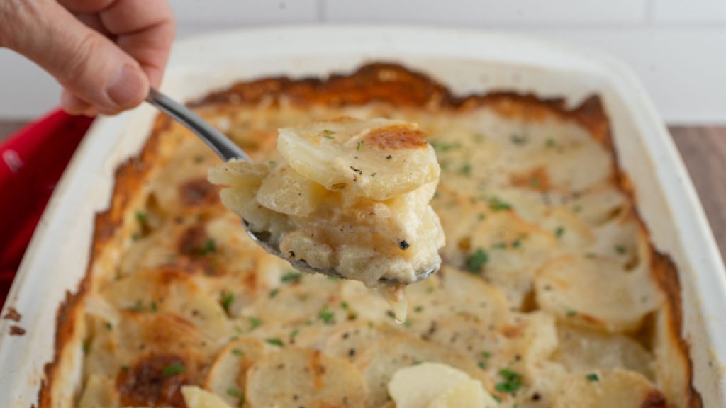casserole dish full of scallop potatoes with a hand holding a scoop out over dish