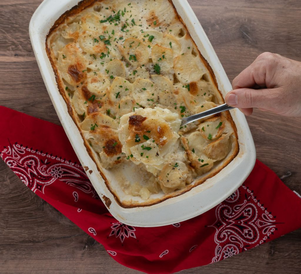 pan of scallop potatoes on a wood background with a red bandana in the corner, hand holding scoop of potatoes out over dish