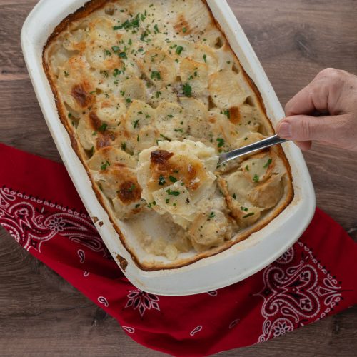 casserole dish full of scallop potatoes with a hand holding a scoop above the dish