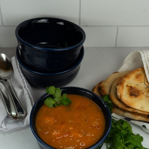 sweet potato curry vegan one pot soup in bowl, extra bowls stacked in back with spoons, naan bread off to the side wrapped in a white towel
