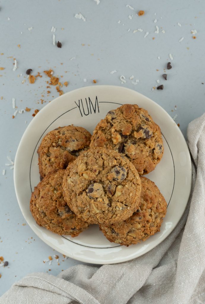 whole wheat granola date chocolate chip cookies on a plate that has the word yum on it sitting on a grey counter top with granola, coconut, and chocolate chips scattered on counter
