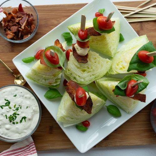Mini wedge salad skewers on white platter with dressing, tomatoes, bacon and mozzarella on the side.