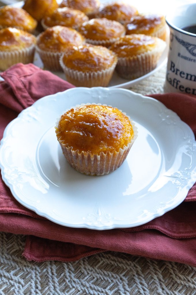 orange kissed muffin on a white plate with plate of muffins in the background with a cup of coffee to the rt top of the photo