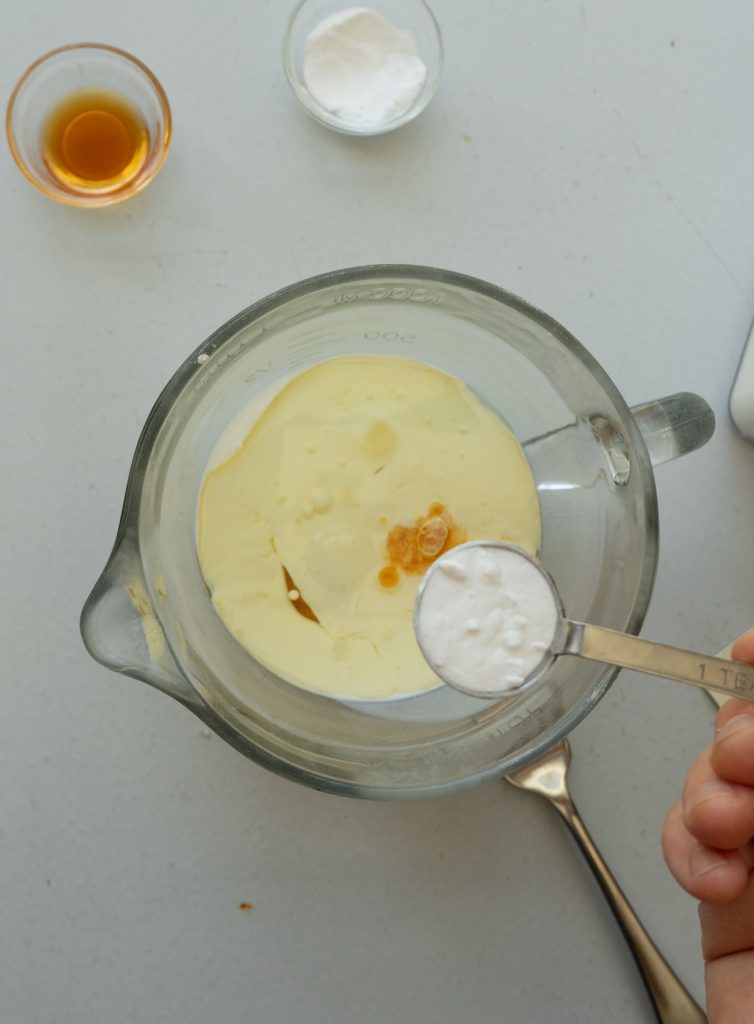wet ingredients in a glass measuring bowl with hand holding a teaspoon of baking soda over the bowl