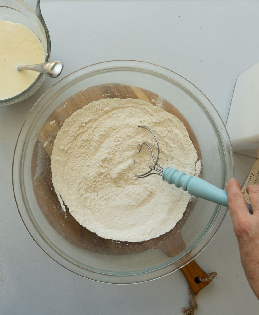 dry ingredients in a glass bowl with a hand coming in from the right side to mix