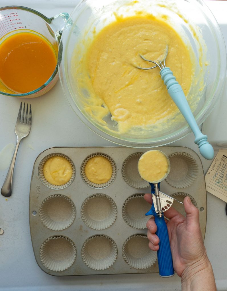scooping muffin batter into prepared muffin tin, bowl of batter is on top to the rt, orange juice reduction is on top of photo to the left