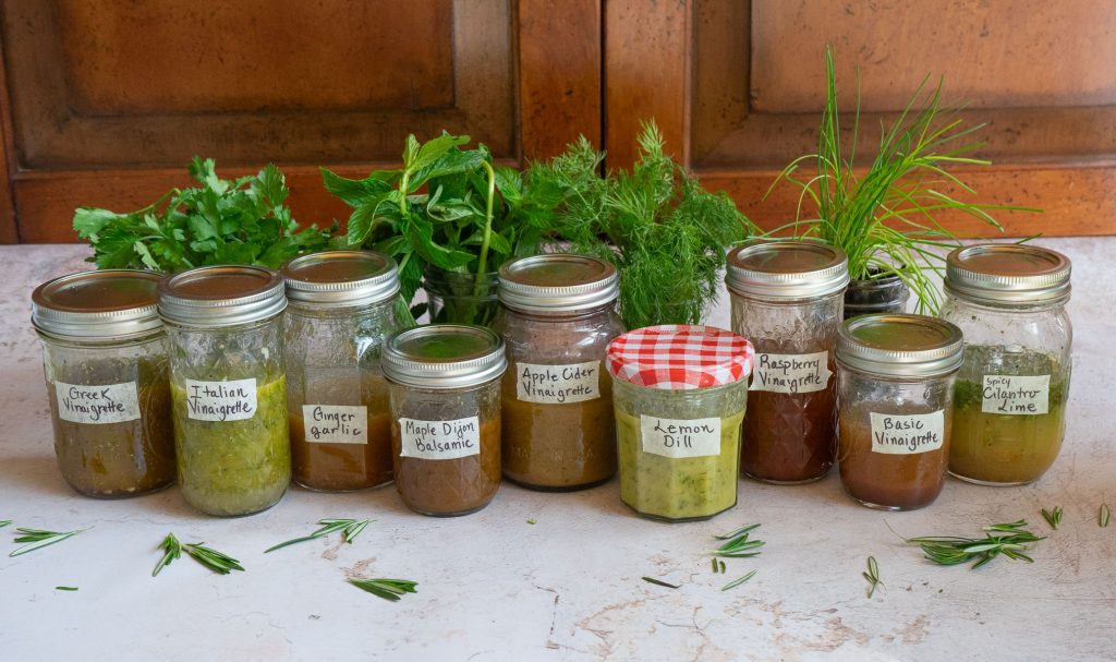 row of homemade vinaigrettes in various glass jars on a gray counter top with wood cupboard in background