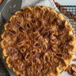 whole pecan pie settin on a cooling rack with pecans strewn on table pie server and knife off to the upper left side of the pie