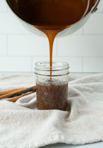 picture of caramel being poured into a glass jar with cinnamon sticks in the background with a tan napkin
