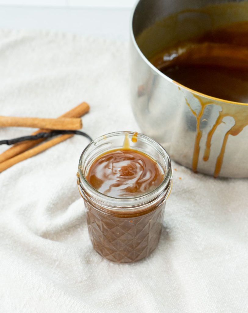 glass jar of caramel sauce sitting on a linen napkin with a vanilla bean and some cinnamon sticks off to the side, pan with caramel sauce dripping down the outside is in the background