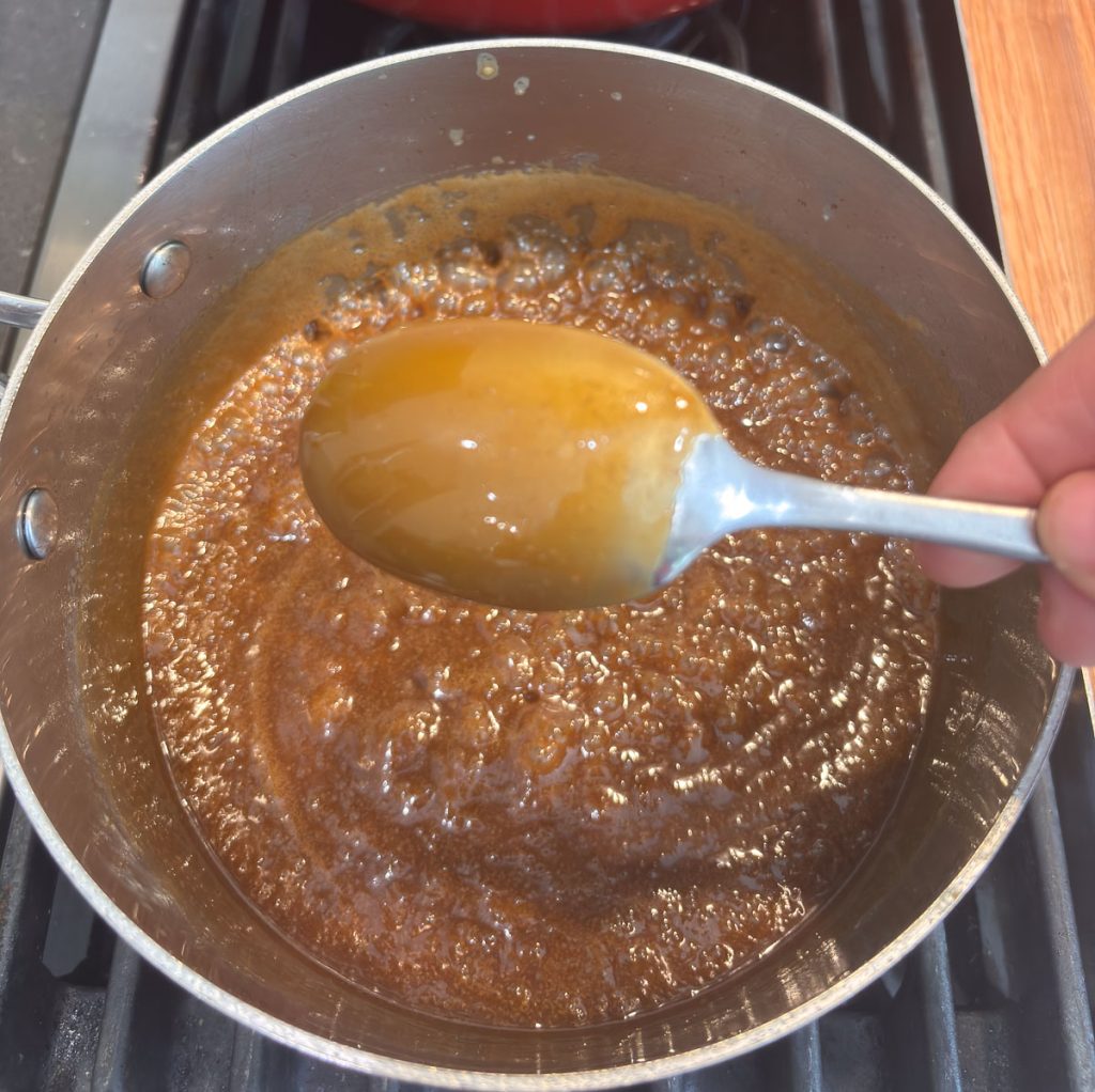 spoon with caramel sauce on it over a pan of bubbling caramel sauce on a stovetop