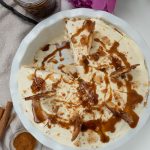 Whole ice cream pie with one slice take out in a white pie dish, with the jar of caramel sauce in the upper left hand corner small dish of cinnamon in bottom left corner flowers also in photo