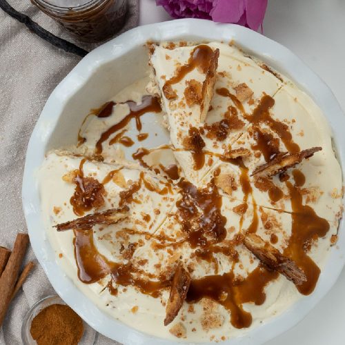 Whole ice cream pie with one slice take out in a white pie dish, with the jar of caramel sauce in the upper left hand corner small dish of cinnamon in bottom left corner flowers also in photo