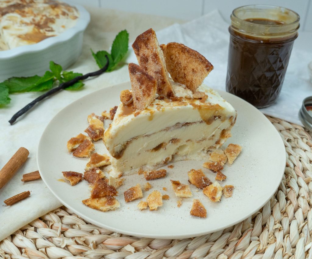 slice of churro ice cream pie on a white plate that is on a raton placemat, with jar of caramel sauce and the whole pie in the background