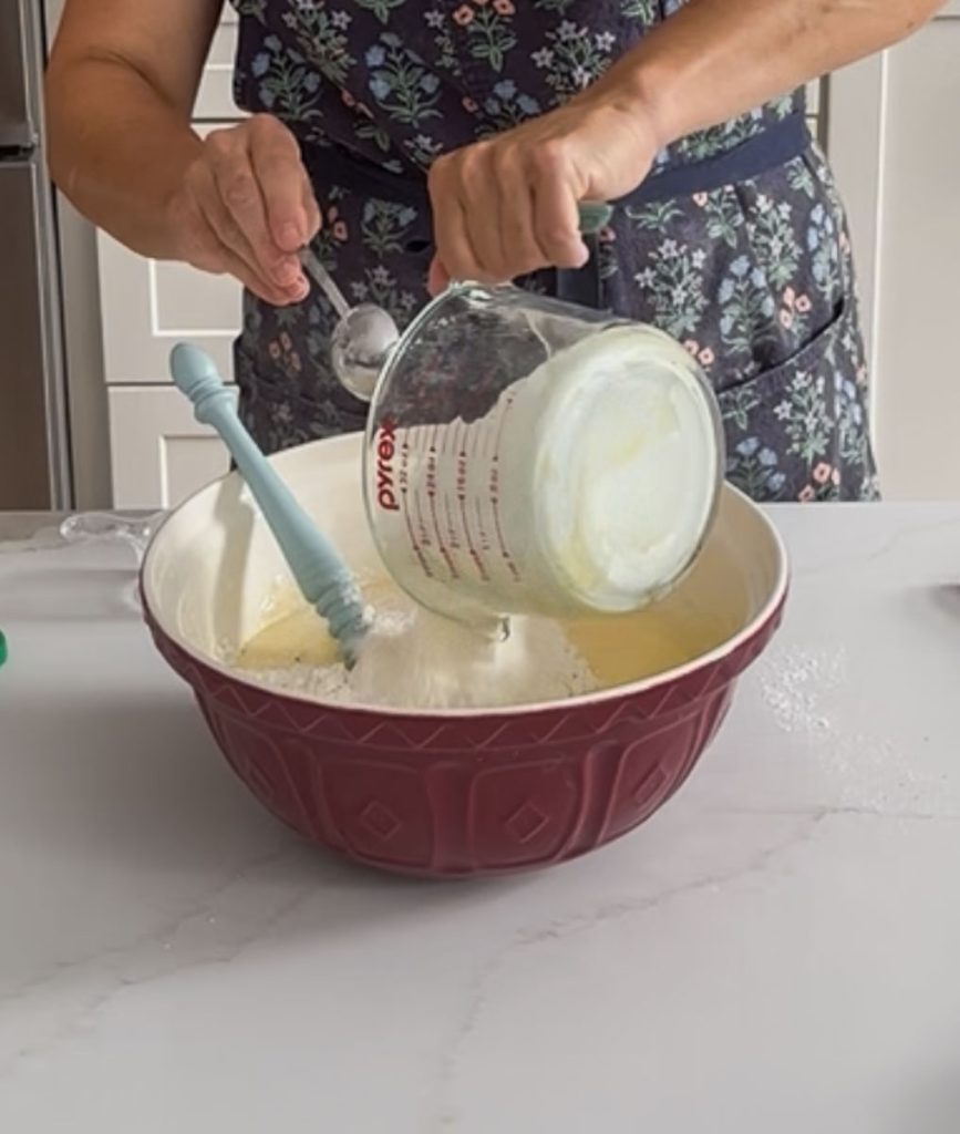 adding dry ingredients to wet ingredients that are in a large red bowl sitting on a white coutner top