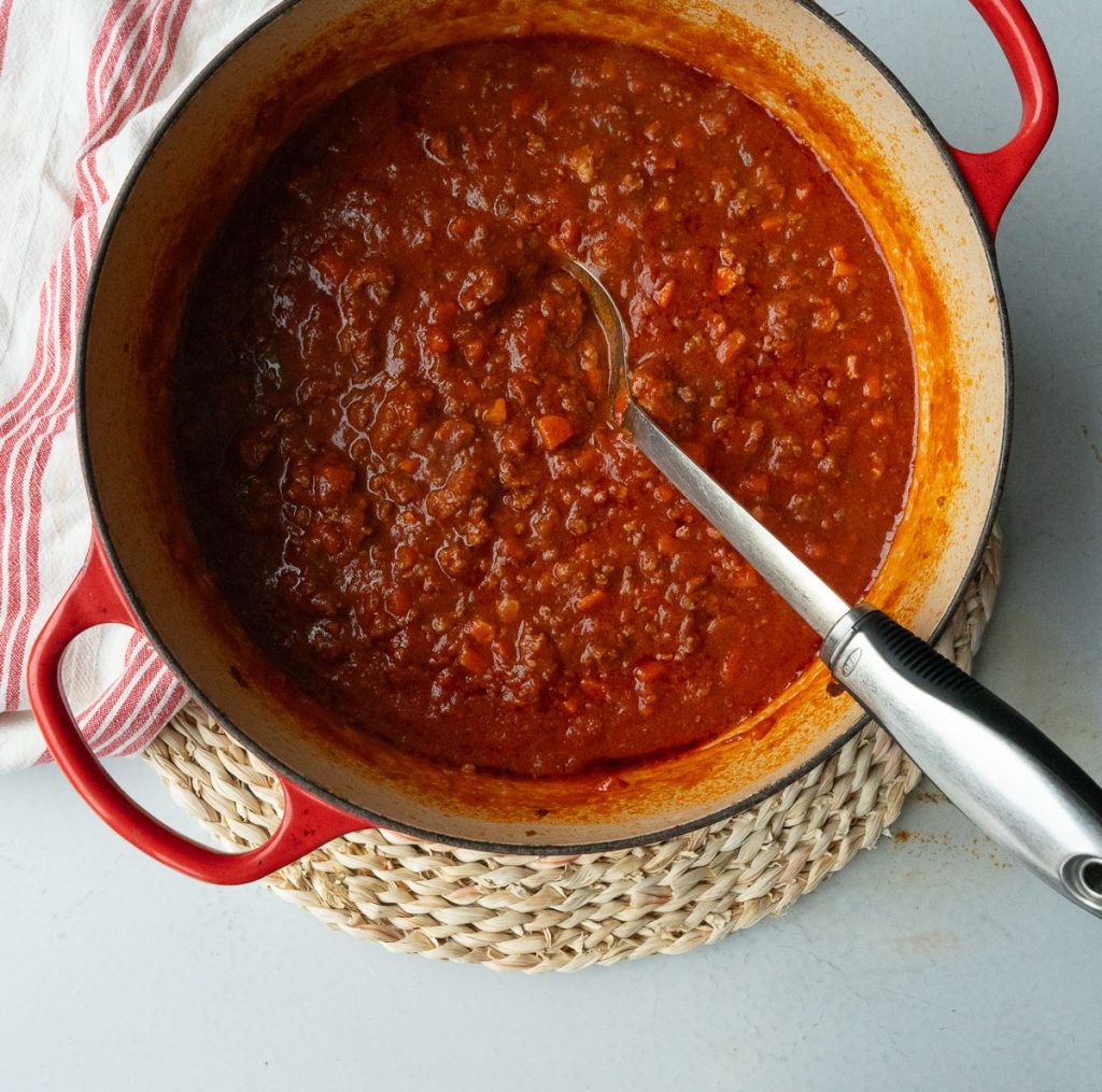 pot of ragu in a red dutch oven that is sitting on a rattan place mat with a spoon in the pot