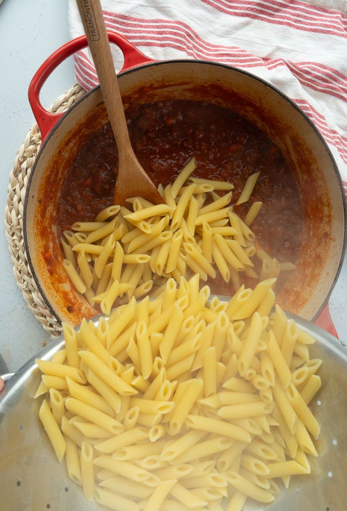 adding hot cooked ziti noodles to pot of ragu sauce, wooden spoon is in pot