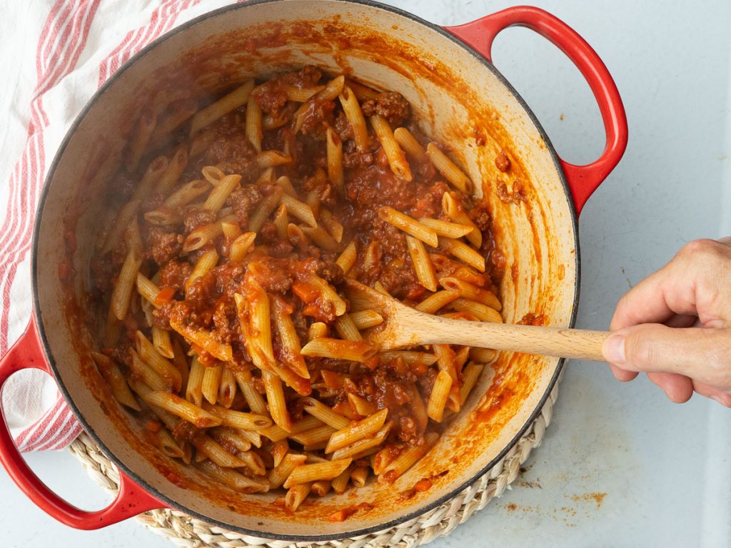 stirring ziti pasta into the ragu sauce in red dutch oven a hand is stirring with a wooden spoon