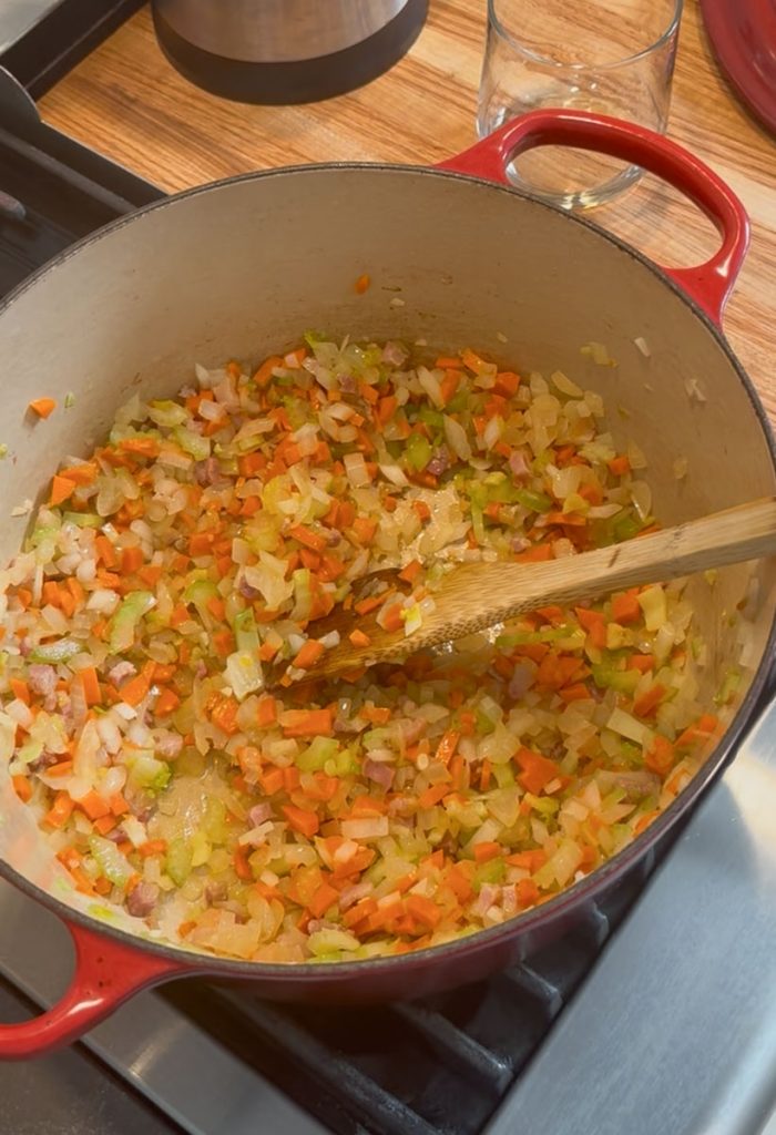 red dutch oven on the stove with onion, carrot, and celery sauting in it with a wooden spoon setting in the pot