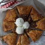 apple crisp galette sliced on baking sheet with three scoops of ice cream in the middle of the galette with a red gingham cloth in the top of photo with forks on top of the cloth