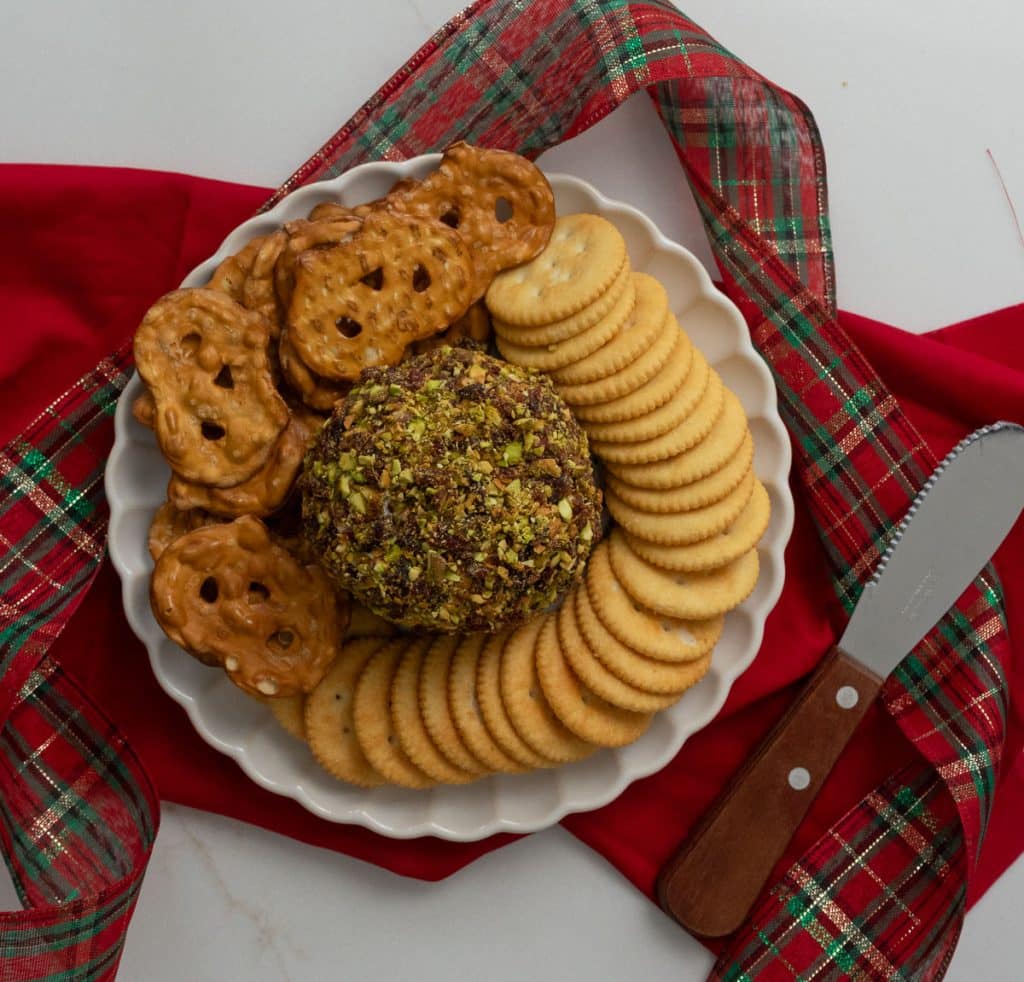 white plate with cheese ball in the middle surrounded by ritz crackers and pretzel crisps with holiday ribbon and a red cloth in the back ground