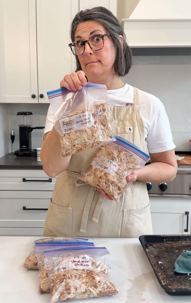person in a tan apron and white t-shirt holding up zip-loc bags of shredded chicken 