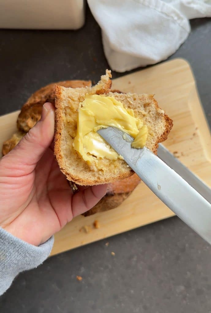 hands holding a slice of irish soda bread and spreading butter on the slice over a wood cutting board on a black countertop