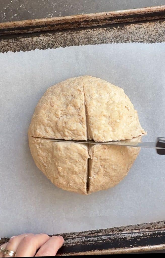 irish soda bread dough ball on a parchment lined baking sheet being cut crosswise with a knife